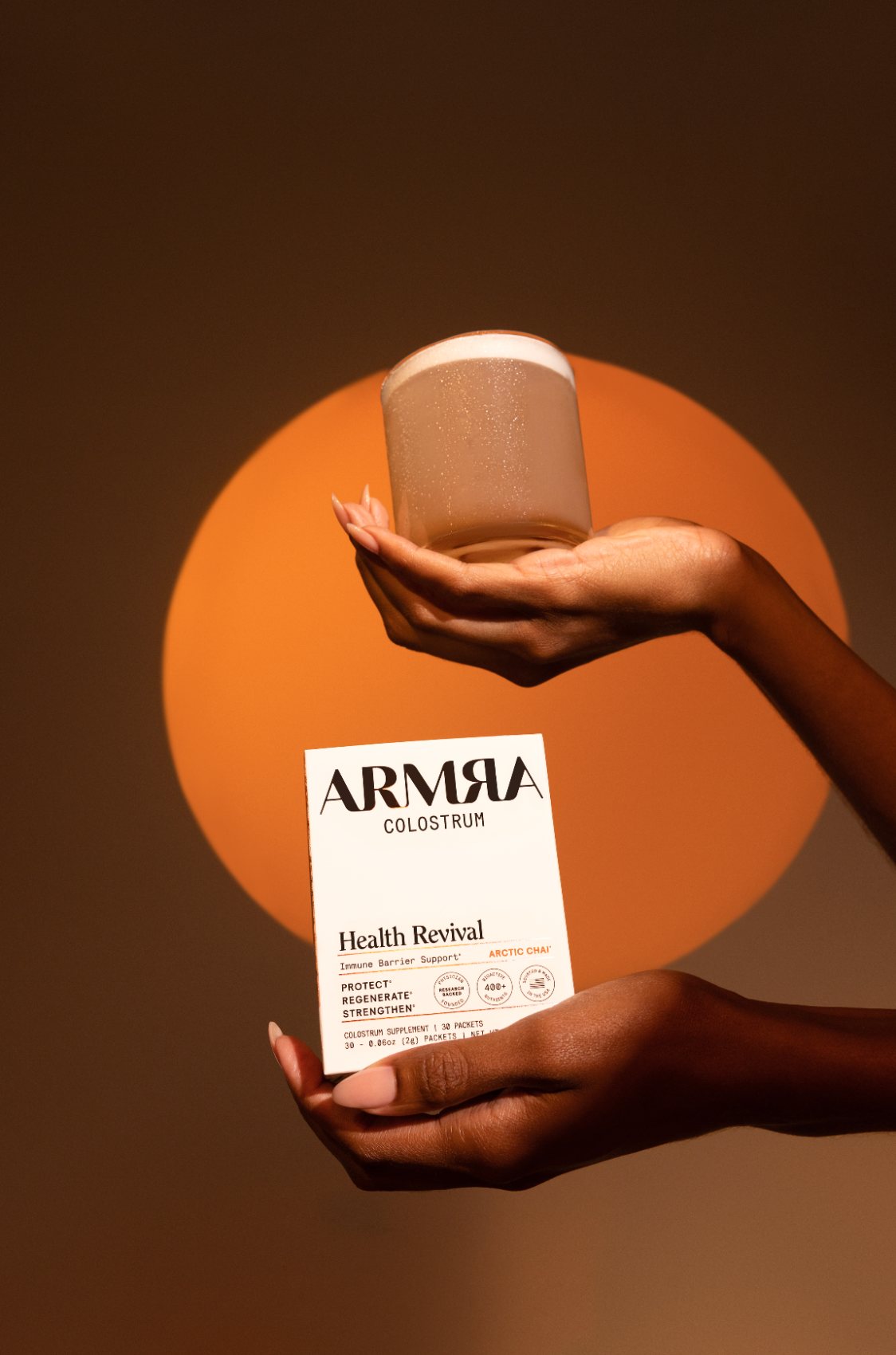 Hands holding a glass and an ARMRA Colostrum box in front of an orange disc