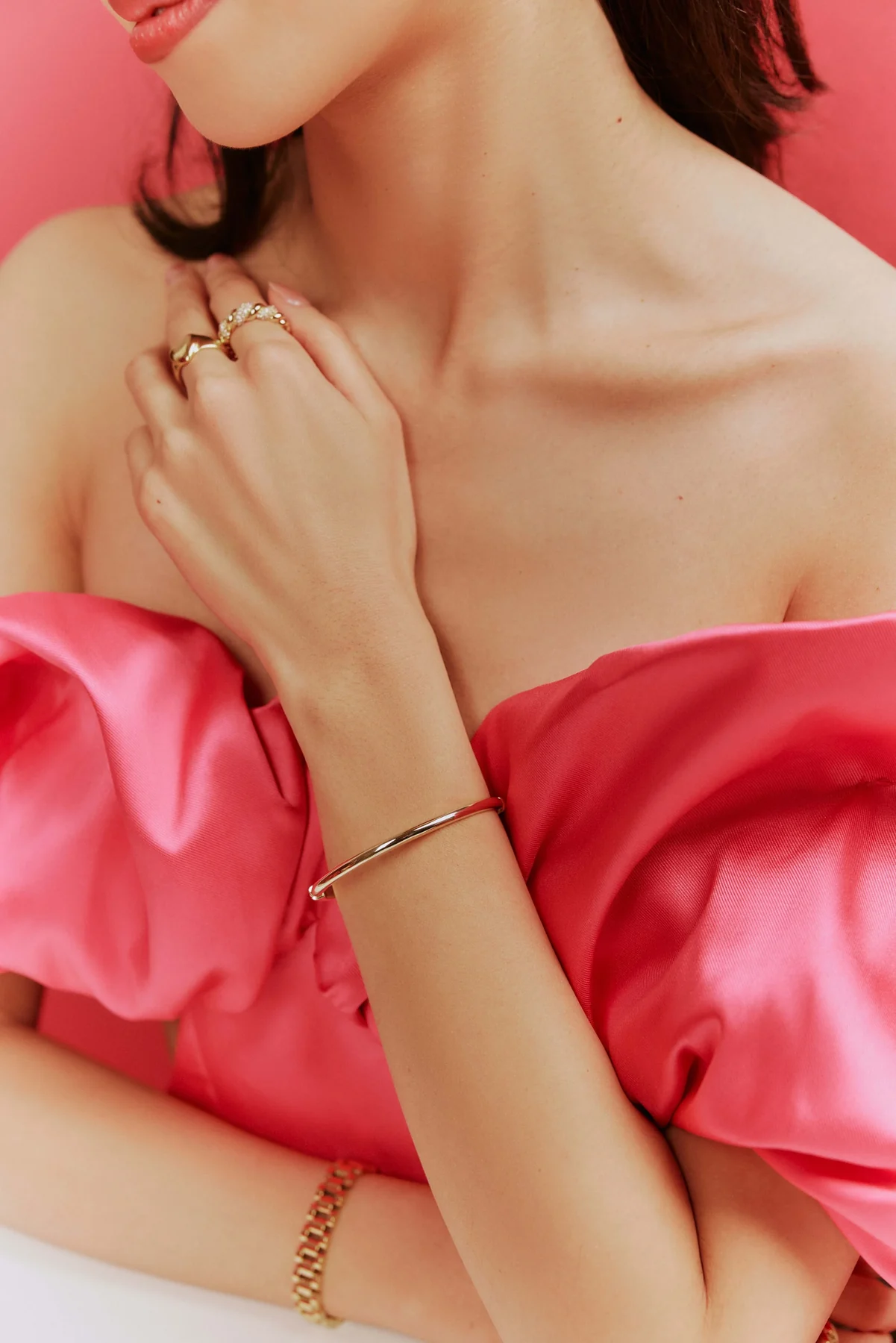 Model in pink satin touching her collarbone, wearing gold rings and a bangle