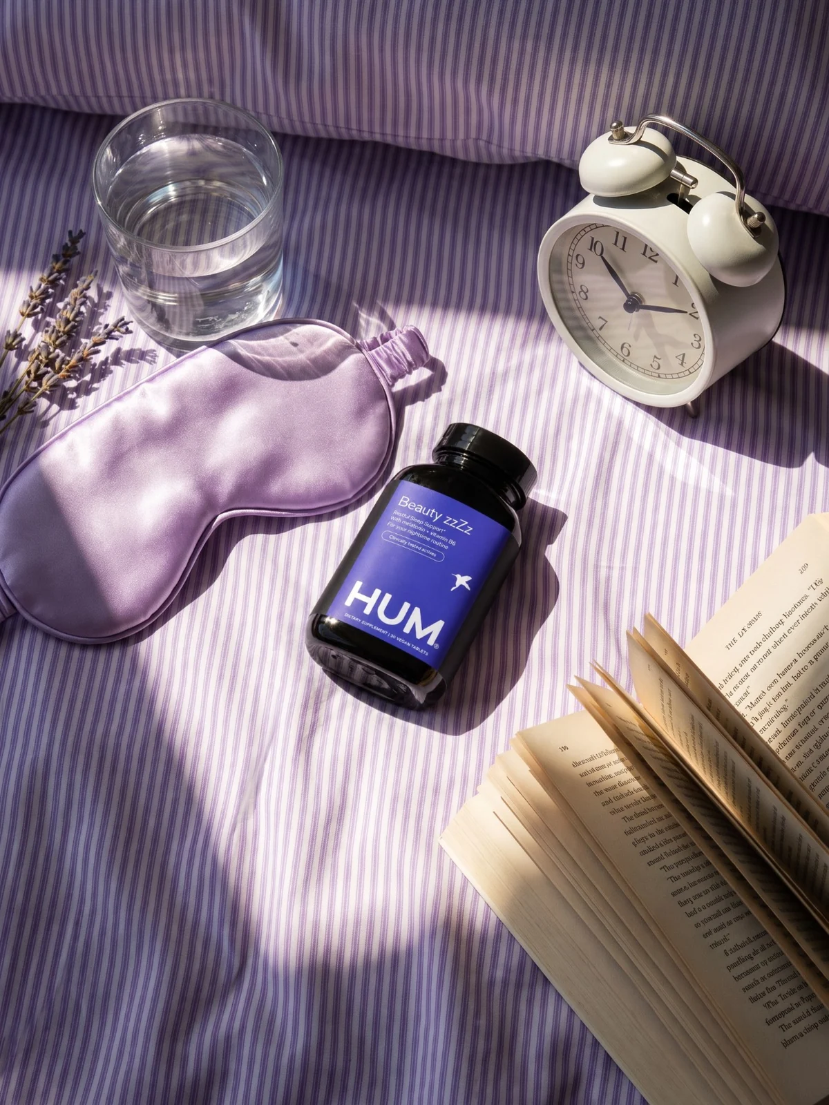 Beauty zzZz bottle on violet striped sheets with a sleep mask, alarm clock, water and an open book