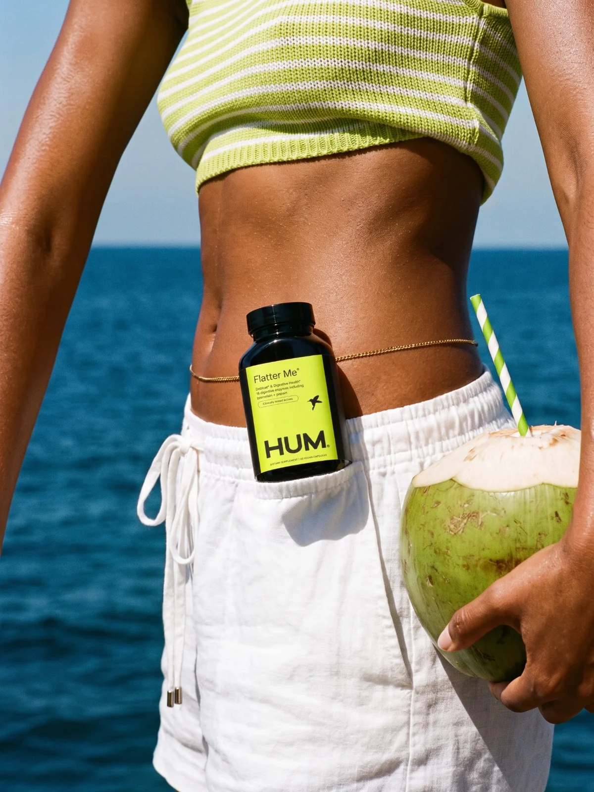 Close crop of a waist by the sea, a Flatter Me bottle tucked in white shorts and a coconut in hand